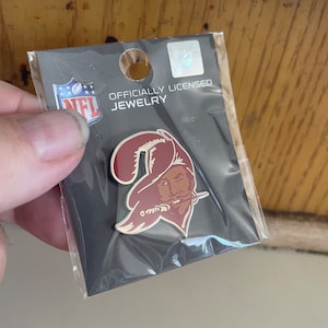 May include: A pin featuring the Tampa Bay Buccaneers logo, a pirate with a red hat and a white outline, on a black card. The card is labeled "OFFICIALLY LICENSED JEWELRY" and includes the NFL logo. The pin is in a clear plastic package.