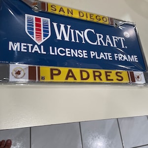 May include: A blue metal license plate frame with the text "SAN DIEGO WINCRAFT METAL LICENSE PLATE FRAME" and a red, white, and blue logo. The bottom of the frame is yellow with the text "PADRES" in brown letters.