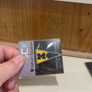 May include: A silver and blue Michigan Wolverines pin on a black and white card with the text "Official Licensed Collectible Pins & Earrings" and "WinCraft Collection".