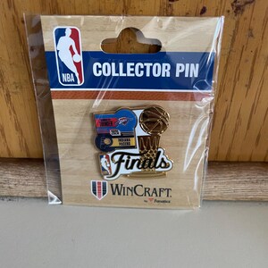 May include: A collector pin featuring NBA and Finals logos, a basketball, and the text "COLLECTOR PIN". The pin is packaged in a clear plastic sleeve and includes the WinCraft logo.