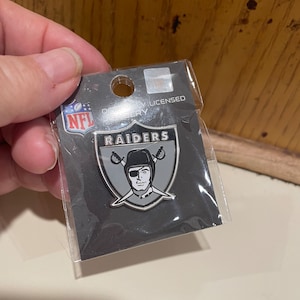 May include: A silver Raiders NFL pin with a black and white logo of a pirate wearing an eye patch and holding crossed swords. The pin is packaged in a clear plastic bag with a black and white card that says "Officially Licensed NFL".