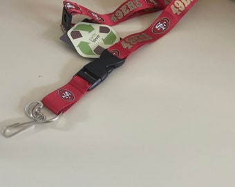 Peut inclure: Lanière rouge et or avec le texte "49ers" répété. La lanière a un clip en plastique noir et un crochet en métal argenté. Une petite étiquette verte avec le texte "earth love" est attachée à la lanière.