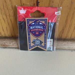 May include: A gold and blue NCAA Men's Basketball National Champions pin with the year 2022 and the text "Final Four New Orleans".