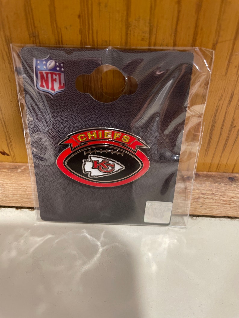 Pu&ograve; includere: Uno spillo da football dei Kansas City Chiefs nero e rosso con il logo della squadra e la parola "CHIEFS" in lettere gialle su uno stendardo rosso.