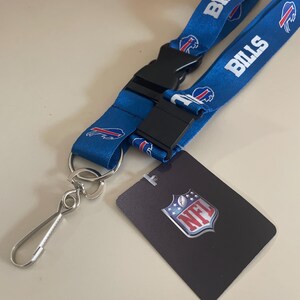 Op de afbeelding: Een blauwe lanyard met het Buffalo Bills logo en het woord "Bills" herhaald op de band. De lanyard heeft een zwarte plastic clip en een zilveren metalen haak. De lanyard wordt geleverd met een kleine kaart met het NFL-logo.
