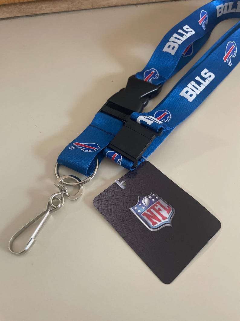 Op de afbeelding: Een blauwe lanyard met het Buffalo Bills logo en het woord "Bills" herhaald op de band. De lanyard heeft een zwarte plastic clip en een zilveren metalen haak. De lanyard wordt geleverd met een kleine kaart met het NFL-logo.