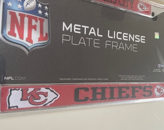 May include: A metal license plate frame with a red and white Kansas City Chiefs logo. The frame is decorated with the NFL logo and the words "Metal License Plate Frame".