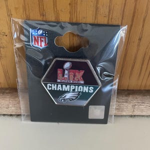May include: A silver and green hexagonal pin with the text "NFL Super Bowl Champions" and a Philadelphia Eagles logo. The pin is packaged in a clear plastic bag with a black backing card.