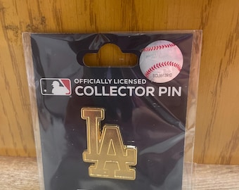 May include: A gold-colored LA logo collector pin, officially licensed by MLB, is displayed on a black background. The pin is in a clear plastic package, with a baseball graphic and the WinCraft logo.