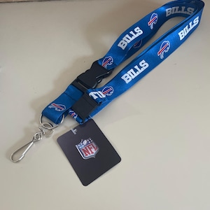 Op de afbeelding: Een blauwe lanyard met het Buffalo Bills logo en het woord "Bills" dat meerdere keren wordt herhaald. De lanyard heeft een zwarte plastic clip en een zilveren metalen haak. Een klein zwart label met het NFL-logo is inbegrepen.