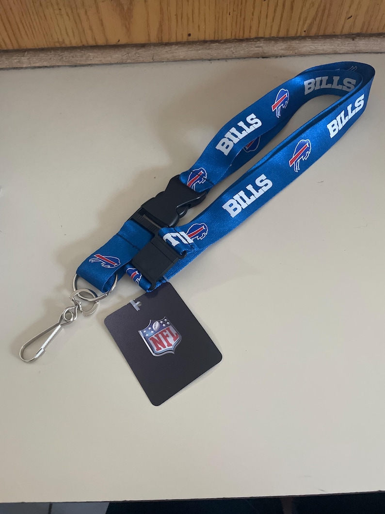 Op de afbeelding: Een blauwe lanyard met het Buffalo Bills logo en het woord "Bills" dat meerdere keren wordt herhaald. De lanyard heeft een zwarte plastic clip en een zilveren metalen haak. Een klein zwart label met het NFL-logo is inbegrepen.