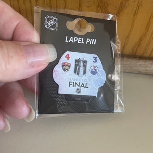 May include: A black and white lapel pin featuring the Stanley Cup Final logo with the team logos for the Florida Panthers and the Edmonton Oilers. The pin has the team scores, 4 and 3, and the text "Stanley Cup Final".
