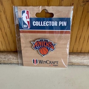 May include: A New York Knicks collector pin featuring the team logo with an orange basketball and blue and orange lettering. The pin is attached to a branded cardboard backing card with the NBA logo, the text COLLECTOR PIN, and WINCRAFT.