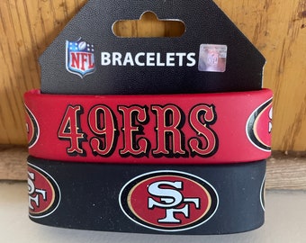 Puede incluir: Dos pulseras de silicona con el logotipo de los San Francisco 49ers. La pulsera superior es roja con las palabras "49ers" en blanco. La pulsera inferior es negra con el logotipo de los 49ers en rojo y blanco.