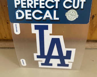 May include: A white and blue "LA" decal on a clear background. The decal is packaged in a plastic sleeve with a header that reads "Perfect Cut Decal" and "Proudly Made in the USA".