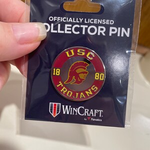 May include: Officially licensed USC Trojans collector pin with a red, gold, and white design. The pin features a helmet with a Spartan warrior and the text "USC Trojans" around the helmet. The pin also has the numbers "18" and "80" on either side of the helmet. The pin is made by WinCraft by Fanatics.