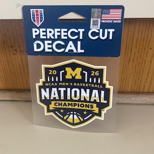 May include: A Perfect Cut Decal featuring the text "2026 NCAA Men's Basketball National Champions". The decal has a blue and yellow color scheme with the American flag and the words "Proudly Made".