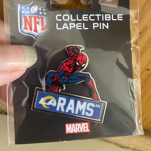 May include: Collectible lapel pin featuring Spider-Man in a dynamic pose, above a blue sign that reads "RAMS AVE." The pin is set against a black background and includes the NFL and Marvel logos.
