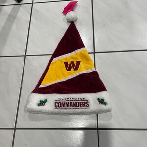 May include: A burgundy and gold Washington Commanders Christmas hat with a white trim. The hat features the team's logo and the word "Commanders" in white lettering. A white pom-pom sits at the top.