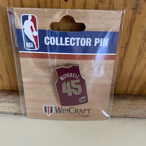 May include: A red and gold basketball jersey pin with the name "Mitchell" and the number "45" on the back. The pin is on a white card with the words "COLLECTOR PIN" and "WINCRAFT" on it.