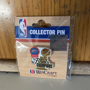 May include: A collector pin in a clear plastic package. The pin features the NBA logo, the text "COLLECTOR PIN", and the words "Finals Champions" with a basketball design. The pin also has the text "2025" and the WinCraft logo.