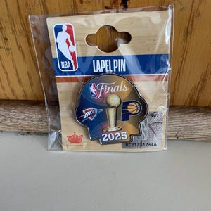 May include: A lapel pin featuring the NBA logo and the text "Finals 2025." The pin has a basketball and trophy design, with OKC branding. The pin is packaged in a clear plastic sleeve.