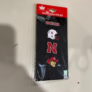 May include: A set of three collectible pins featuring the University of Nebraska Cornhuskers logo. The pins are red and white and feature a helmet, a letter N, and a cartoon mascot. The packaging is black with the words "Go Big Red" and "Collectible Pin Set".