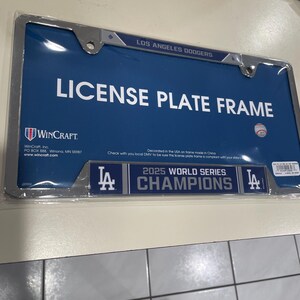 May include: A blue license plate frame with the text "LOS ANGELES DODGERS" at the top. The frame reads "LICENSE PLATE FRAME" in large white letters. The bottom of the frame says "2025 WORLD SERIES CHAMPIONS".