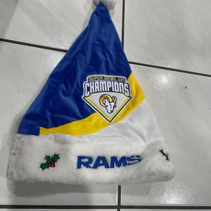 May include: Blue and white Santa hat with a white pom-pom and trim. The hat features the text "SUPER BOWL LVI CHAMPIONS" and the word "RAMS" in blue, along with a team logo and holly accents.