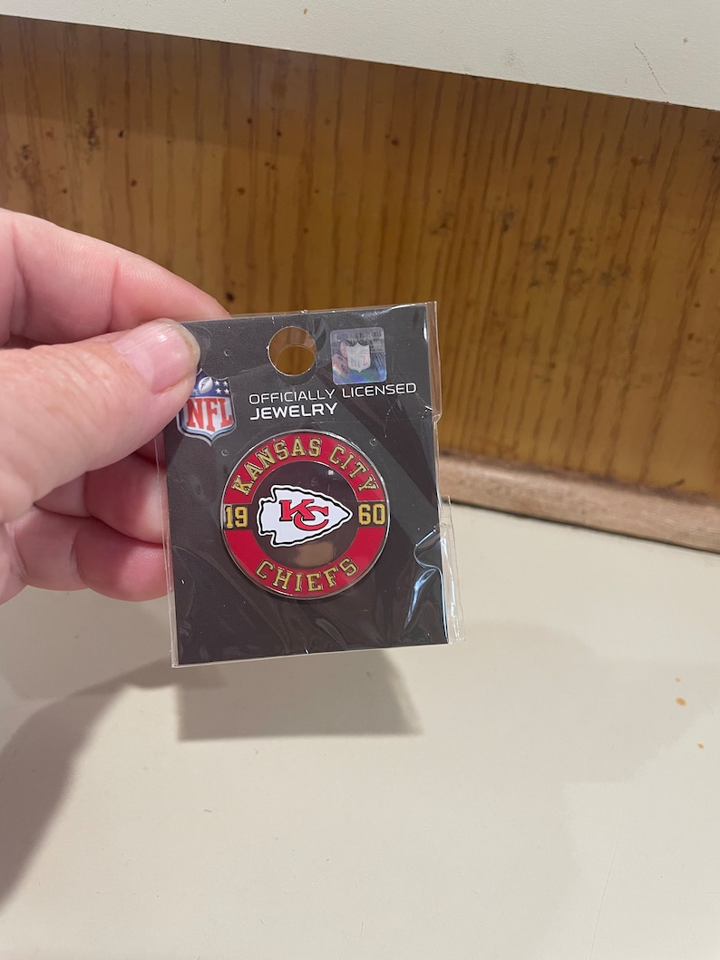 May include: Officially licensed NFL jewelry pin featuring the Kansas City Chiefs logo with the years 19 and 60 on either side of the logo. The pin is red, white, and gold.