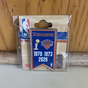 May include: A rectangular metal pin featuring a blue banner design with the text 3X NBA CHAMPIONS, the New York Knicks logo, a trophy graphic, and the years 1970, 1973, and 2026. The pin is mounted on a cardboard backing with an NBA logo in a plastic sleeve.