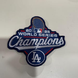 May include: A blue and white embroidered patch with the text "2025 World Series Champions" and the Los Angeles Dodgers logo. The patch has a baseball theme and is likely a collectible item.