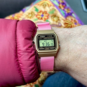 Puede incluir: Un reloj digital Casio F-91W dorado con una correa de nailon rosa. La esfera del reloj muestra la hora, la fecha y el d&iacute;a de la semana. El reloj es resistente al agua.