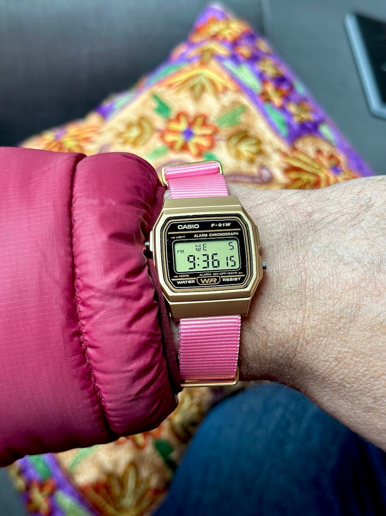 Puede incluir: Un reloj digital Casio F-91W dorado con una correa de nailon rosa. La esfera del reloj muestra la hora, la fecha y el d&iacute;a de la semana. El reloj es resistente al agua.