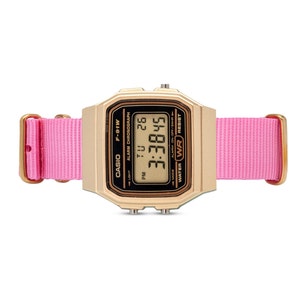 Puede incluir: Un reloj digital Casio de color dorado con una correa de nailon rosa. La esfera del reloj muestra la hora, la fecha y el d&iacute;a de la semana. El reloj es resistente al agua.