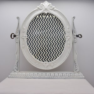 May include: White vanity mirror with an oval frame and a black and white chevron patterned backing. The mirror has ornate details and a decorative base.