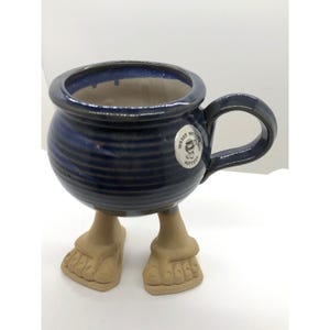 May include: A round, dark blue glazed ceramic mug with horizontal ridges and a single handle. The mug is supported by two sculpted, light brown ceramic feet wearing sandals. A circular white sticker on the side features the text Muddy Waters Pottery.