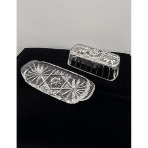 May include: A clear glass butter dish consisting of a rectangular base with rounded ends and a matching lid. Both pieces feature a pressed glass pattern of radiating starbursts and geometric shapes, creating a textured, reflective surface.