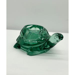 May include: A translucent emerald green glass turtle figurine featuring a hollow shell opening at the top. The turtle is depicted in a crawling position with a smooth, molded surface, defined head, and four legs, crafted from clear green glass material.