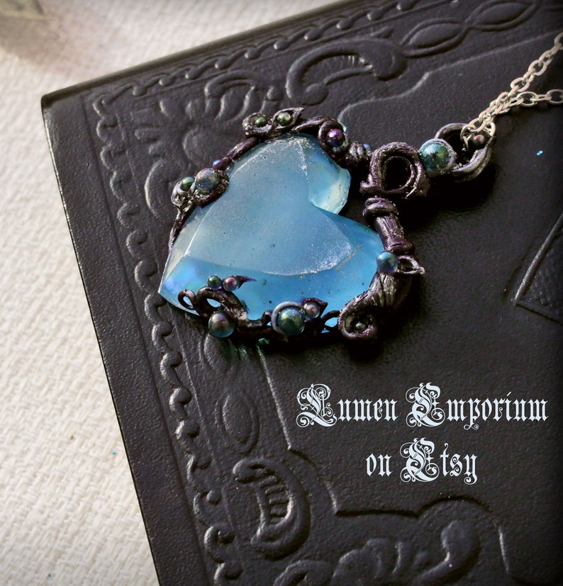 May include: A blue glass heart pendant with black and silver wire wrapped around it. The pendant is adorned with small black and iridescent beads. The pendant is hanging from a silver chain.