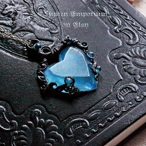May include: A blue heart-shaped pendant necklace with black filigree detailing and dark blue beads. The necklace is on a black leather-bound journal.