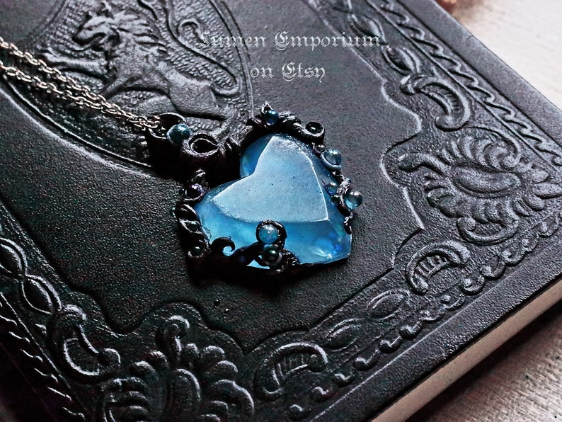 May include: A blue heart-shaped pendant necklace with black filigree detailing and dark blue beads. The necklace is on a black leather-bound journal.