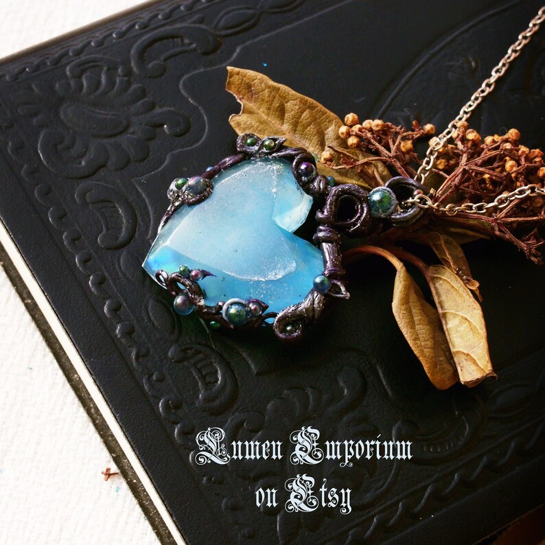 May include: A blue heart-shaped pendant necklace with a silver and black wire wrapped around it. The pendant is surrounded by small black and green beads. The necklace is on a black background.