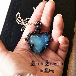 May include: A blue heart-shaped pendant necklace with a black wire frame and iridescent beads. The pendant is hanging from a silver chain.