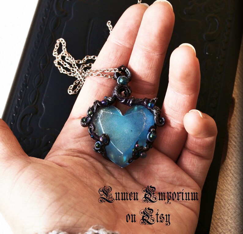 May include: A blue heart-shaped pendant necklace with a black wire frame and iridescent beads. The pendant is hanging from a silver chain.