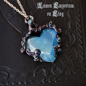 May include: A blue heart-shaped pendant necklace with intricate silver and black details. The pendant is surrounded by small silver and black beads. The necklace is on a black background.