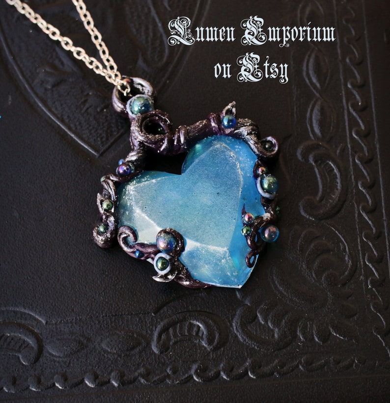May include: A blue heart-shaped pendant necklace with intricate silver and black details. The pendant is surrounded by small silver and black beads. The necklace is on a black background.