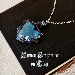 May include: A blue heart-shaped pendant necklace with a silver chain. The pendant is made of resin and has a sparkly finish. It is surrounded by black wire and small iridescent beads. The necklace is displayed on a black leather-bound journal with the text "Lumen Emporium on Etsy".