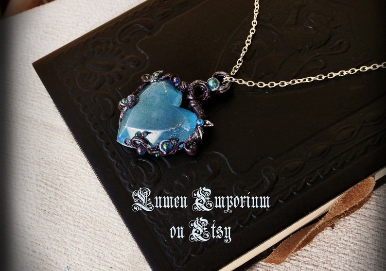 May include: A blue heart-shaped pendant necklace with a silver chain. The pendant is made of resin and has a sparkly finish. It is surrounded by black wire and small iridescent beads. The necklace is displayed on a black leather-bound journal with the text "Lumen Emporium on Etsy".