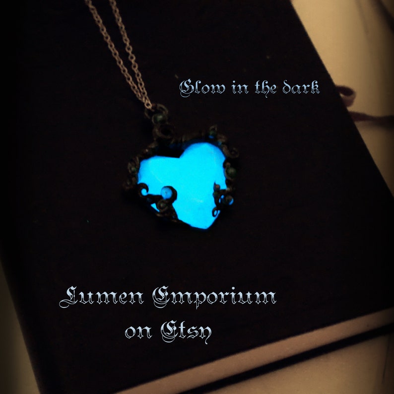 May include: A glowing blue heart-shaped pendant necklace with a black metal filigree design. The necklace is on a black surface and the text "Glow in the dark" and "Lumen Emporium on Etsy" is visible.